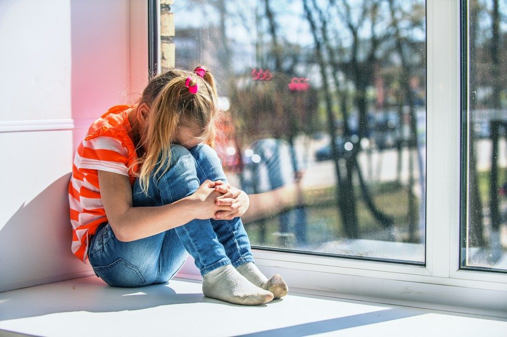 Little girl in orange blouse sits on a window sill, crying