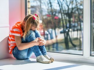 Little girl in orange blouse sits on a window sill, crying