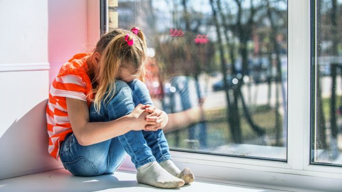 Little girl in orange blouse sits on a window sill, crying